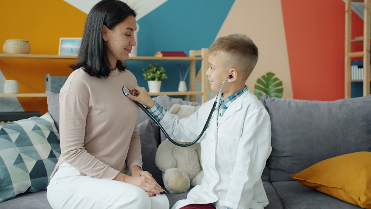 Little boy in white gown is playing with mom examining her with stethoscope while woman is smiling and breathing enjoying game with cute child.
